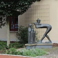 Bronzestatue eines Spielmanns mit Flöte und zuhörendem Kind vor einer beige Gebäude.