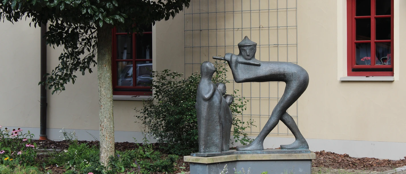 Bronzestatue eines Spielmanns mit Flöte und zuhörendem Kind vor einer beige Gebäude.