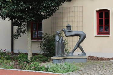 Skulptur "Der Spielmann" Bronzestatue eines Spielmanns mit Flöte und zuhörendem Kind vor einer beige Gebäude.