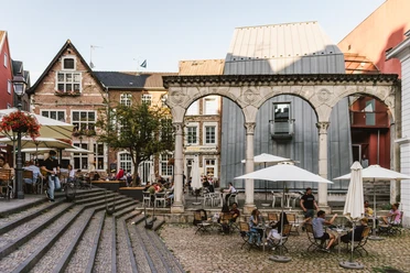 Belebter Hof in Aachen mit historischen Gebäuden, einem römischen Torbogen und Café-Terrassen, wo Menschen entspannt sitzen.