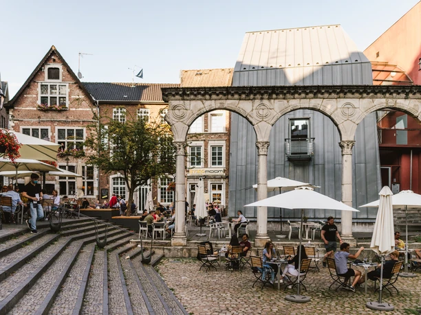 Hof Aachen Belebter Hof in Aachen mit historischen Gebäuden, einem römischen Torbogen und Café-Terrassen, wo Menschen entspannt sitzen.