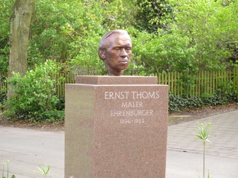 Eine Bronzebüste von Ernst Thoms steht auf einem steinernen Sockel in einer grünen Parkanlage.