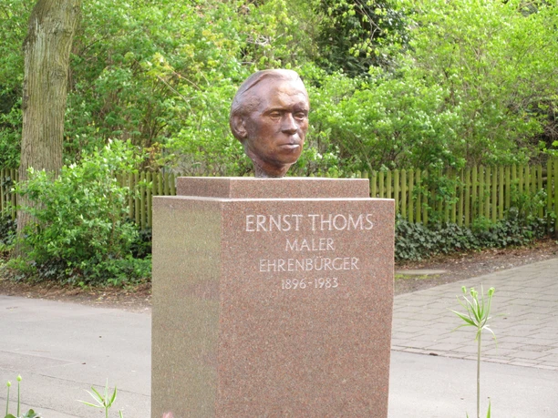 Eine Bronzebüste von Ernst Thoms steht auf einem steinernen Sockel in einer grünen Parkanlage.
