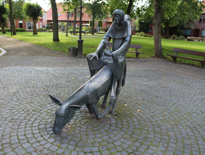 Skulptur eines Mönchs auf einem bückenden Esel inmitten eines gepflasterten, von Bäumen umgebenen Platzes.