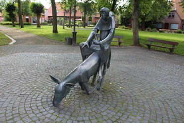 Skulptur Esel und Mönch Skulptur eines Mönchs auf einem bückenden Esel inmitten eines gepflasterten, von Bäumen umgebenen Platzes.