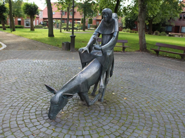 Skulptur Esel und Mönch Skulptur eines Mönchs auf einem bückenden Esel inmitten eines gepflasterten, von Bäumen umgebenen Platzes.