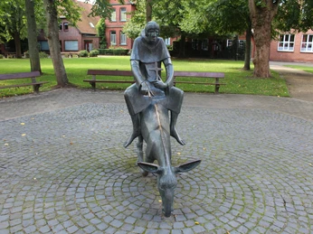 Skulptur Esel und Mönch Skulptur von einem Mönch auf einem Esel, umgeben von Bäumen und roten Backsteinbauten.