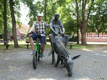 Skulptur Esel und Mönch Ein Mann auf einem Fahrrad posiert neben einer Bronzeskulptur eines Mönchs auf einem Esel im Park.