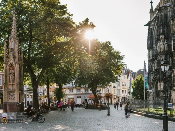 Gepflasterter Platz mit einer historischen Säule und großen Bäumen in der Mitte, durch die die Abendsonne scheint. Rechts sieht man einen Teil des Aachener Doms.