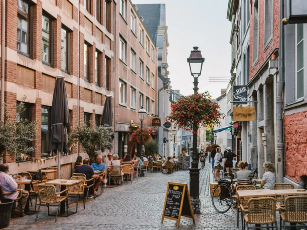 Gepflasterte Gasse zwischen bunten Häuserreihen und historischen Straßenlaternen, die mit Blumenkästen geschmückt sind. Vor den Häusern stehen Sitzgruppen von Restaurants oder Cafés, an denen Personen sitzen.