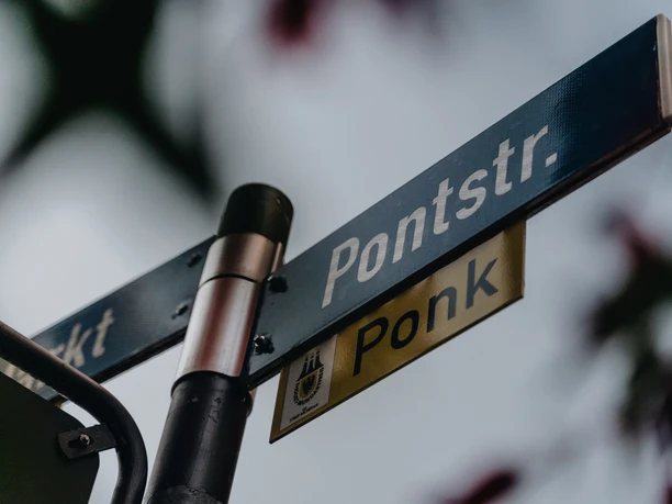 Pontstraße Aachen Schild Wegweiser mit mehreren Straßenschildern, darunter eins mit der Aufschrift "Pontstraße" und darunter "Ponk", dem Straßennamen in Aachener Dialekt.