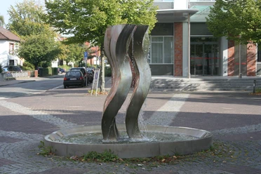 Drei geschwungene Stahlskulpturen thronen in einem kreisförmigen Brunnen auf einem gepflasterten Platz.