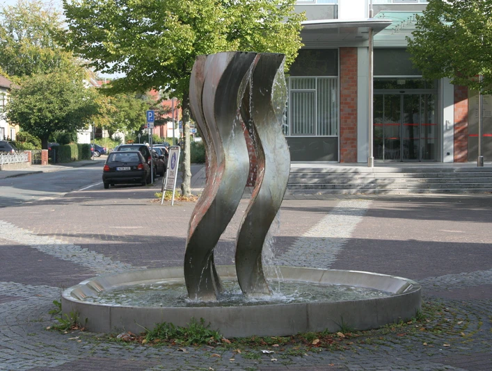 Drei geschwungene Stahlskulpturen thronen in einem kreisförmigen Brunnen auf einem gepflasterten Platz.