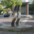 Die Drei Grazien Drei geschwungene Stahlskulpturen thronen in einem kreisförmigen Brunnen auf einem gepflasterten Platz.