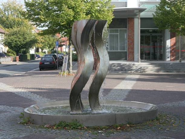 Die Drei Grazien Drei geschwungene Stahlskulpturen thronen in einem kreisförmigen Brunnen auf einem gepflasterten Platz.