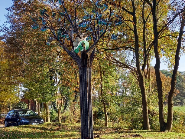 Eiche mit Schild-Lieblingsort Die Metallskulptur einer stilisierten Eiche steht an einem Rastplatz, umrahmt von herbstlich gefärbten Bäumen und einer mit Laub bedeckten Wiese im Vordergrund. Im Hintergrund sind weitere Bäume und ein blauer Himmel sichtbar.