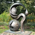 Eine Bronze-Skulptur mit abstrakten Formen steht vor einer blühenden Gartenkulisse.