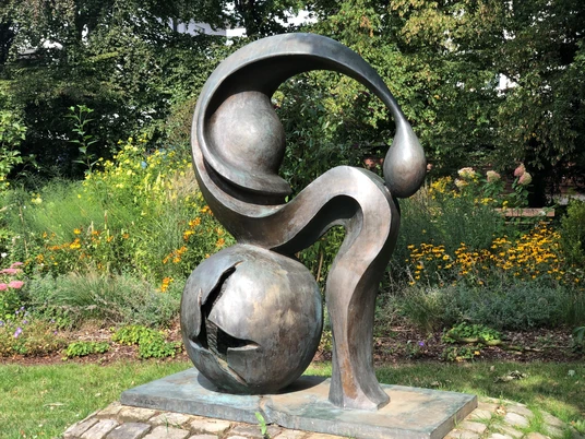 Eine Bronze-Skulptur mit abstrakten Formen steht vor einer blühenden Gartenkulisse.