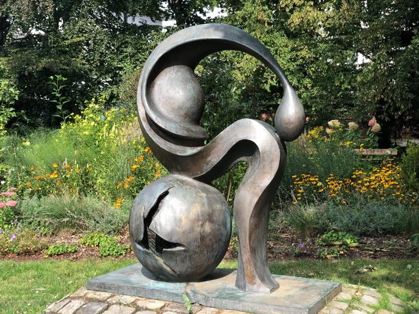 Eine Bronze-Skulptur mit abstrakten Formen steht vor einer blühenden Gartenkulisse.