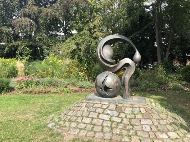 Eine Skulptur aus Metall, die organisch geschwungene Formen zeigt, steht auf einem gepflasterten Sockel im Grünen.
