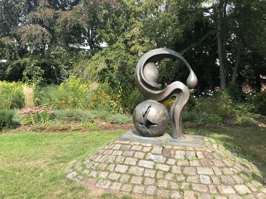 Madonna von Tschernobyl Eine Skulptur aus Metall, die organisch geschwungene Formen zeigt, steht auf einem gepflasterten Sockel im Grünen.