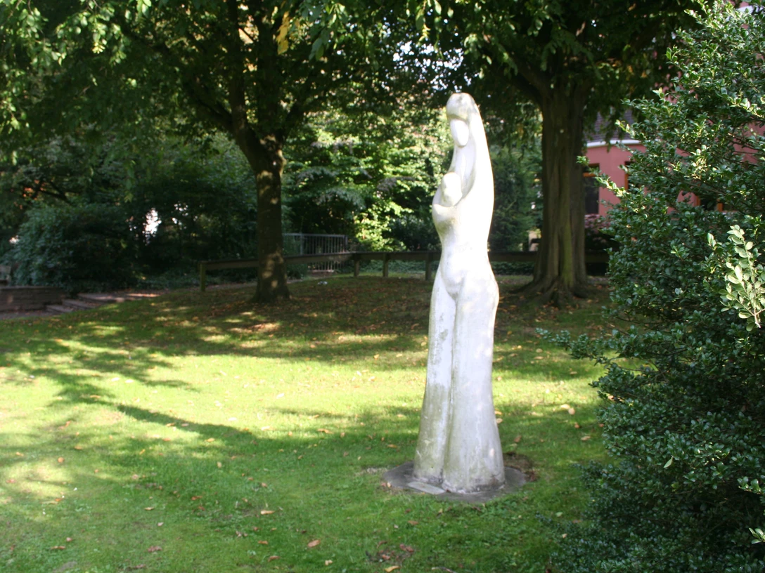 Die Skulptur einer stehenden Figur in einem sonnigen Park, umgeben von Bäumen und grünem Gras.
