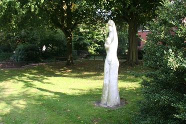 Marion + Weser Die Skulptur einer stehenden Figur in einem sonnigen Park, umgeben von Bäumen und grünem Gras.