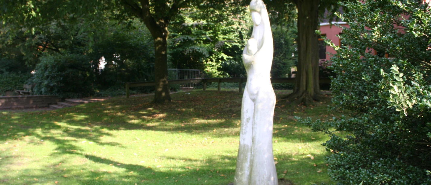 Marion + Weser Die Skulptur einer stehenden Figur in einem sonnigen Park, umgeben von Bäumen und grünem Gras.