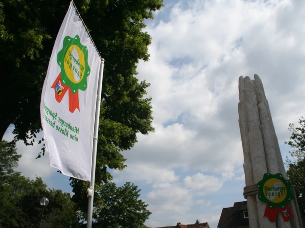 Nienburger Spargelfigur Spargelfigur in Nienburg; hohe weiße Skulptur neben Flagge im Park; blauer Himmel mit Wolken.