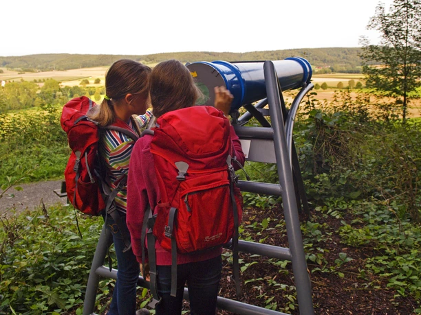 Zwei Mädchen mit roten Rucksäcken betrachten die Landschaft durch ein Sphäriskop in einer grünen Umgebung.