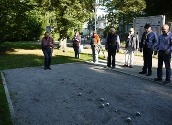 Boule-Bahn am Kurpark Das Bild zeigt mehrere Menschen, die an der Boulebahn stehen und gerade spielen. Das Wetter ist sonnig, und der Kurpark erblüht in voller Pracht.