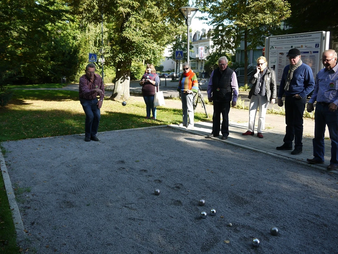 Boule-Bahn am Kurpark Das Bild zeigt mehrere Menschen, die an der Boulebahn stehen und gerade spielen. Das Wetter ist sonnig, und der Kurpark erblüht in voller Pracht.