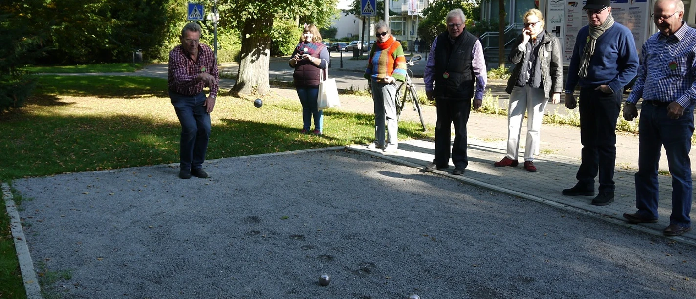 Boule-Bahn am Kurpark Das Bild zeigt mehrere Menschen, die an der Boulebahn stehen und gerade spielen. Das Wetter ist sonnig, und der Kurpark erblüht in voller Pracht.