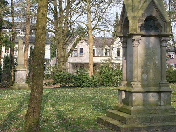 Zwei historische Denkmäler aus Stein stehen auf einem grünen Rasen im Rathauspark umgeben von Bäumen.