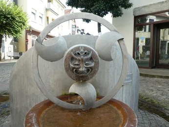 Ein moderner Brunnen mit einem skulpturalen Gesicht aus Metall, umgeben von eleganten Bögen.