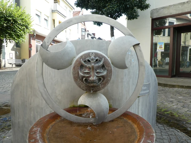 Wasserspielbrunnen Faszination Wasser Ein moderner Brunnen mit einem skulpturalen Gesicht aus Metall, umgeben von eleganten Bögen.