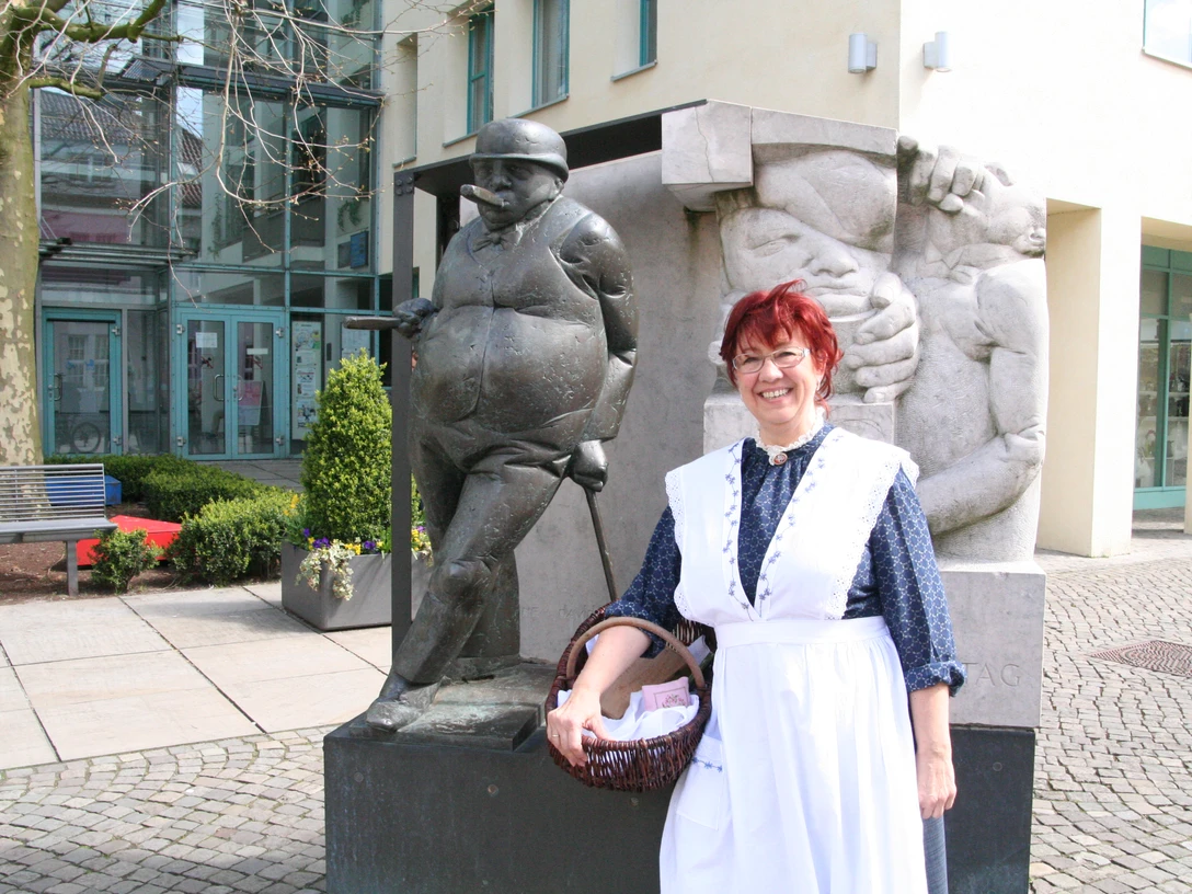 Frau in traditioneller Kleidung steht lächelnd neben einer Bronzeskulptur in einem belebten Innenhof.