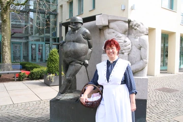 Kinderführung mit Anna - Frau vom Zigarrenmacher Frau in traditioneller Kleidung steht lächelnd neben einer Bronzeskulptur in einem belebten Innenhof.