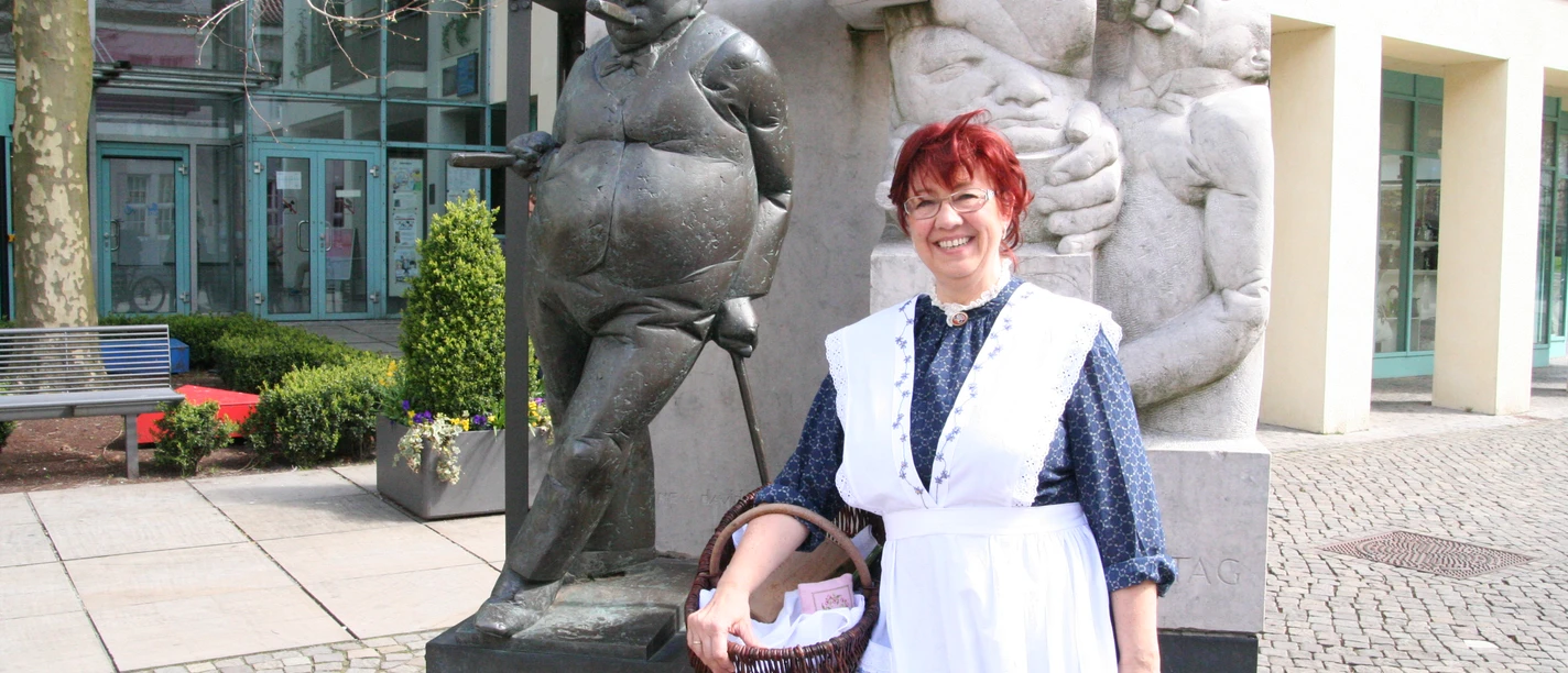 Kinderführung mit Anna - Frau vom Zigarrenmacher Frau in traditioneller Kleidung steht lächelnd neben einer Bronzeskulptur in einem belebten Innenhof.