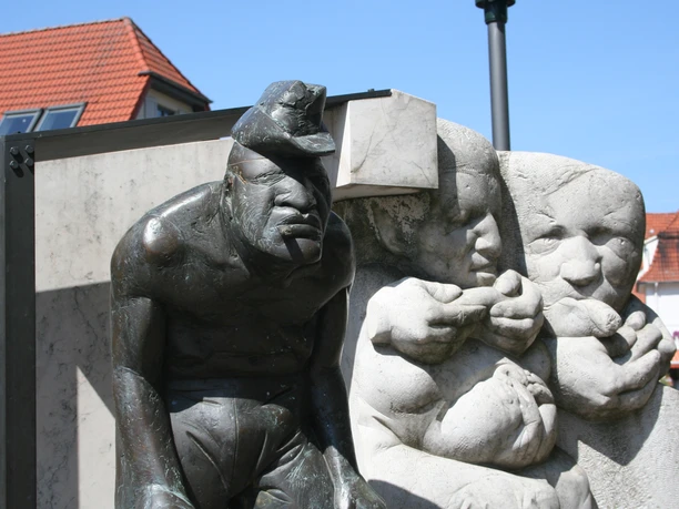 Bronzeskulptur eines Zigarrenmachers und Kindern in rustikaler Straßenszene mit roten Ziegeldächern.