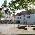 Historische Häuser mit Fachwerkdetails umgeben einen offenen Platz mit Bäumen, Sitzbänken und Brunnen.