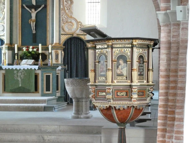 Ein kunstvoll verzierter hölzerner Kanzelkorb mit biblischen Szenen, umrahmt von Altarbereich und Taufbecken.