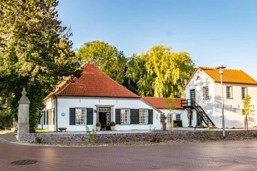 Alte Drostei Weißes historisches Gebäude mit roten Ziegeldächern, hohen Fenstern und Turm vor grünem Baumbestand.