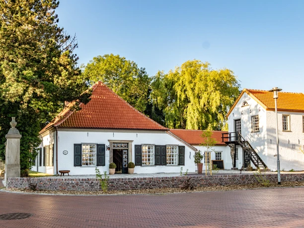 Alte Drostei Weißes historisches Gebäude mit roten Ziegeldächern, hohen Fenstern und Turm vor grünem Baumbestand.