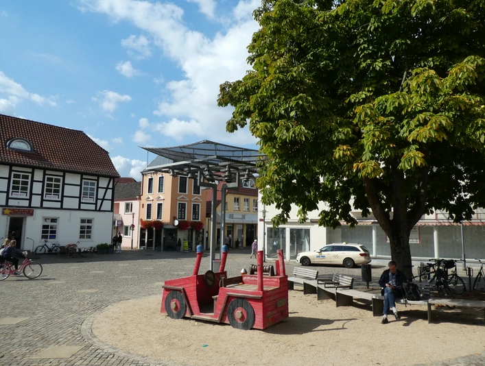 Auf einem sonnigen Marktplatz in Achim sitzt eine Person neben einem roten Holzspielzeugauto unter einem Baum.