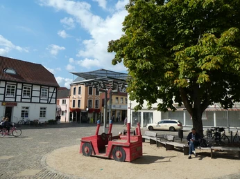 Achimer Glockenspiel Auf einem sonnigen Marktplatz in Achim sitzt eine Person neben einem roten Holzspielzeugauto unter einem Baum.