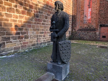 Widukind Steinernes Standbild eines mittelalterlichen Ritters mit Schwert und Schild vor einer Backsteinmauer.