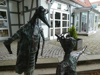 Zwei metallische Skulpturen mit menschlichen Zügen stehen vor einem Fachwerkgebäude mit Kieselsteinen.