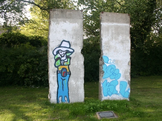 Zwei Segmente der Berliner Mauer mit Graffiti eines Cowboys auf einer grünen Wiese, umgeben von Bäumen.