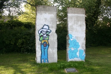 Berliner Mauer Zwei Segmente der Berliner Mauer mit Graffiti eines Cowboys auf einer grünen Wiese, umgeben von Bäumen.